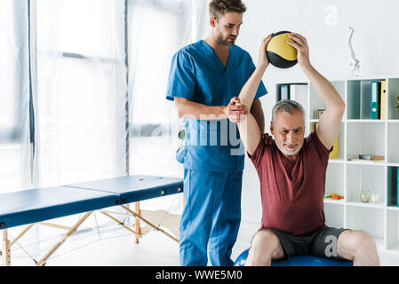 Bello medico in piedi vicino all uomo maturo lavorando sulla sfera di fitness Foto Stock