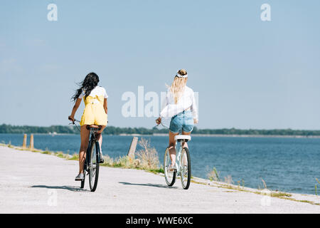 Vista posteriore della bionda e bruna ragazze equitazione biciclette vicino al fiume in estate Foto Stock