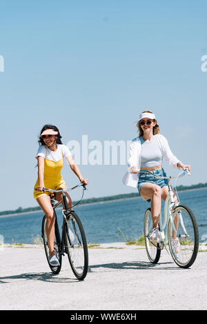 Sorridente bionda e bruna ragazze equitazione biciclette vicino al fiume in estate Foto Stock