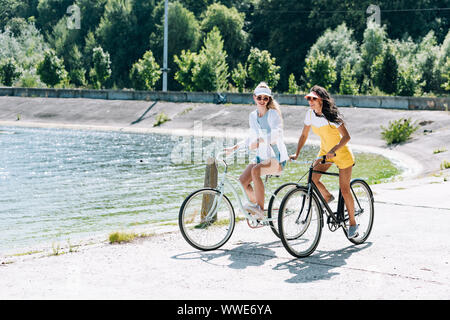 Allegro bionda e bruna ragazze equitazione biciclette vicino al fiume in estate Foto Stock