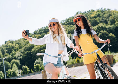 Felice bionda e bruna ragazze equitazione biciclette e tenendo selfie in estate Foto Stock