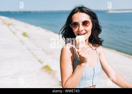 Felice bruna ragazza a mangiare il gelato e equitazione Bicicletta vicino al fiume in estate Foto Stock