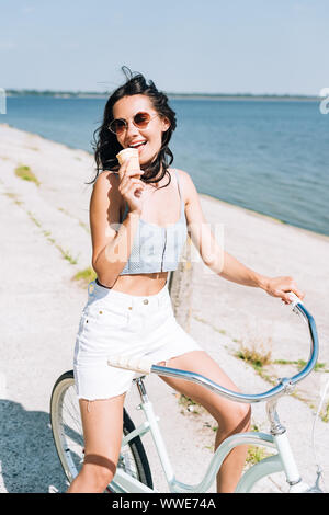 Bruna ragazza a mangiare il gelato e equitazione Bicicletta vicino al fiume in estate Foto Stock