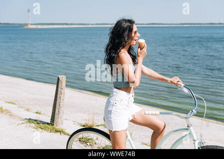 Vista laterale di bruna ragazza a mangiare il gelato e equitazione Bicicletta vicino al fiume in estate Foto Stock