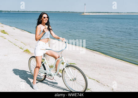 Sorridente ragazza bruna con gelato in Bicicletta Equitazione vicino al fiume in estate Foto Stock