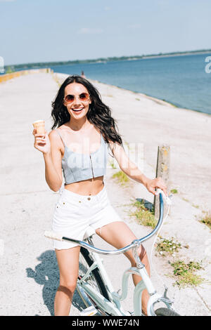 Felice bruna ragazza con gelato in Bicicletta Equitazione vicino al fiume in estate Foto Stock