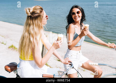 Sorridente bionda e bruna girls riding bikes con gelato vicino al fiume in estate Foto Stock