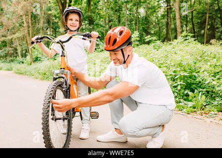 Padre nel casco controllo ruota di bicicletta mentre il figlio in piedi in casco e guardando lontano Foto Stock