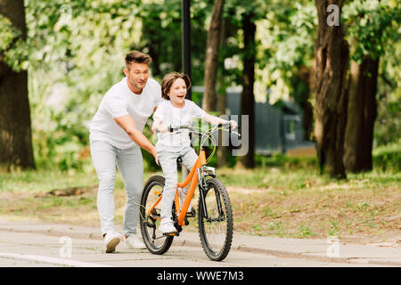 Lunghezza piena vista del padre spingendo bici mentre figlio cacciato in bicicletta Foto Stock