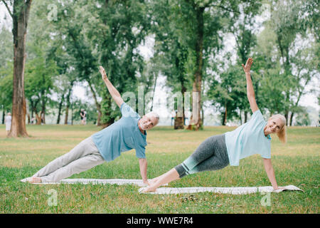 Positivo, uomo maturo e la donna facendo listone laterale esercizio mentre si pratica lo yoga sul prato in posizione di parcheggio Foto Stock