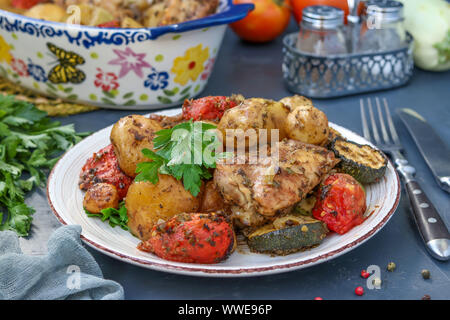 Cosce di pollo al forno con patate, pomodori e zucchine, situato in una piastra e in forma di ceramica, su uno sfondo scuro, orientamento orizzontale Foto Stock