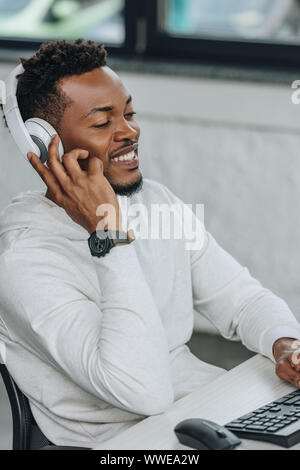 Allegro African American programmer ascoltando musica in cuffia mentre è seduto al posto di lavoro in ufficio Foto Stock