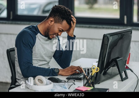 Sconvolto African American programmer tenendo la mano vicino a capo mentre si lavora sul computer in ufficio Foto Stock