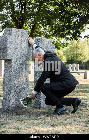 Sconvolto uomo barbuto mettere fiori vicino alla tomba nel cimitero Foto Stock