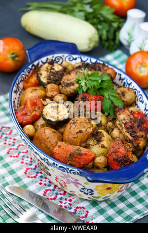 Cosce di pollo al forno con patate, pomodori e zucchine, che si trova in forma di ceramica, su uno sfondo scuro, con orientamento verticale Foto Stock