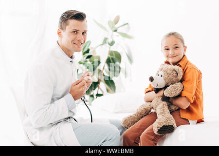 Sorridente pédiatre in camice bianco e capretto con Teddy bear in clinica Foto Stock