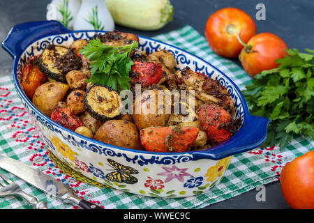 Cosce di pollo al forno con patate, pomodori e zucchine, che si trova in forma di ceramica su uno sfondo scuro, orientamento orizzontale Foto Stock