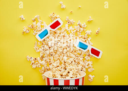 Vista dall'alto di deliziosi popcorn diffusa dalla benna e occhiali 3D su sfondo giallo Foto Stock