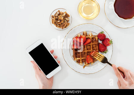 Vista ritagliata della donna di mangiare la colazione e tenendo lo smartphone su bianco Foto Stock