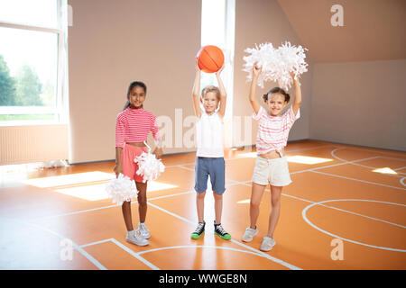 Poco carino cheerleaders in piedi accanto a un compagno di classe con sfera Foto Stock