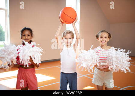 Bel ragazzo carino con sfera in piedi tra cheerleaders Foto Stock