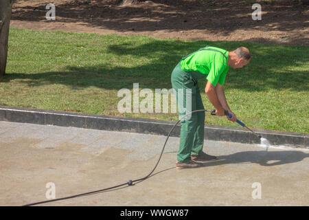 Lavoratore uomo in uniforme lavaggi Street o Park il marciapiede. Servizio municipale della città il processo di pulizia. Guy utilizza acqua apparecchiature di spruzzatura Foto Stock
