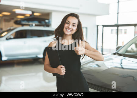 Bella bruna donna manager brochure tenuta in piedi nel centro della concessionaria che mostra come la sign Foto Stock