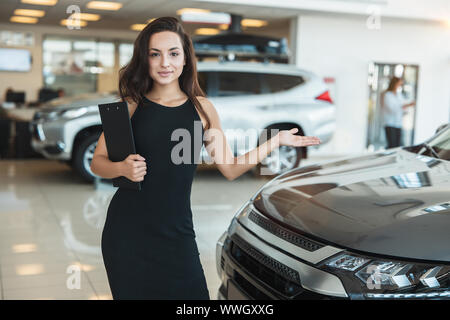 Bella bruna donna manager brochure tenuta in piedi nel centro della concessionaria che mostra la nuova vettura Foto Stock