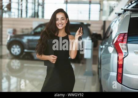 Bella bruna donna manager sorridente azienda brochure in una mano e le chiavi dell'auto in un altro in piedi in concessionaria vicino centro nuovo di zecca SUV Foto Stock