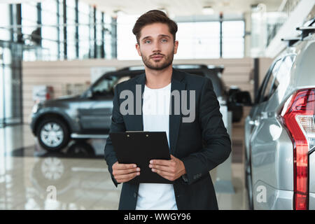 Giovane uomo bello manager sorridente brochure tiene nelle sue mani in piedi in concessionaria vicino centro nuovo di zecca SUV Foto Stock