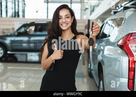 Bella bruna donna manager sorridente tenendo premuto i tasti Auto nella sua mano in piedi in concessionaria vicino centro nuovo di zecca SUV che mostra come Foto Stock