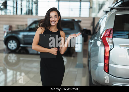 Bella bruna donna manager brochure tenuta in piedi nel centro della concessionaria che mostra auto nuova di zecca Foto Stock