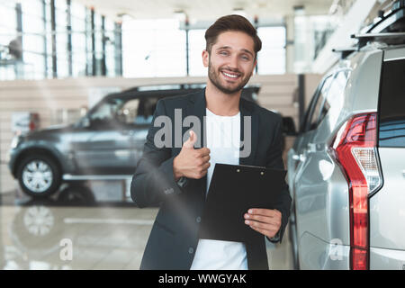 Giovane uomo bello manager sorridente azienda brochure nella sua mano in piedi in concessionaria vicino centro nuovo di zecca SUV che mostra come la sign Foto Stock