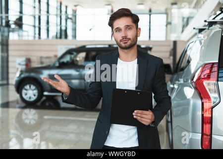 Giovane uomo bello manager sorridente in piedi in concessionaria vicino centro nuovo di zecca SUV con brochure in mano Foto Stock