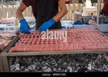 Salsiccia barbecue allineate su un grande grill all'aperto con fumo proveniente dal fuoco sotto. persona in grembiule nero maglietta blu mano tenendo le tenaglie tur Foto Stock