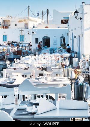 Ristorante all'aria aperta con arredamento bianco contro ancorato di barche e di edifici bianchi nel vecchio porto di Naoussa Grecia in estate giornata di sole Foto Stock