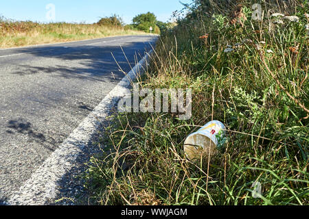 Un basso angolo di visualizzazione di una tazza di cartone che è stato buttato fuori da un veicolo piombo nell'erba sul lato di una strada principale che non abbia alcun traffico su di essa Foto Stock