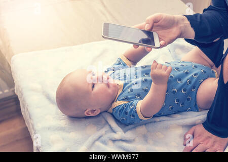 Fotografato neonato sdraiato sul tavolo. La donna è di scattare una foto da smart phone. Il bambino si allunga le mani sul telefono. Tutti i potenziali registrati sono Foto Stock