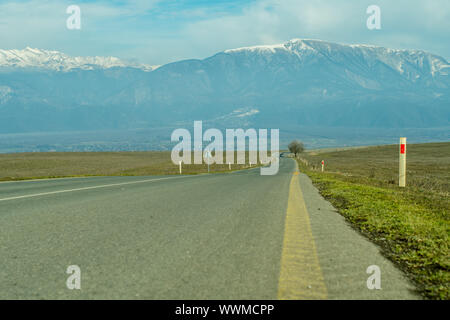 Villaggio di montagna con cime innevate sullo sfondo lungo strada asfaltata in primo piano. Foto Stock