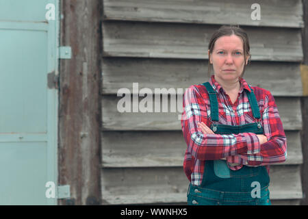 Ritratto di donna agricoltore di fronte al casale capannone. Donna vestita di plid camicia e jeans denim tuta come salariato agricolo con le braccia incrociate Foto Stock