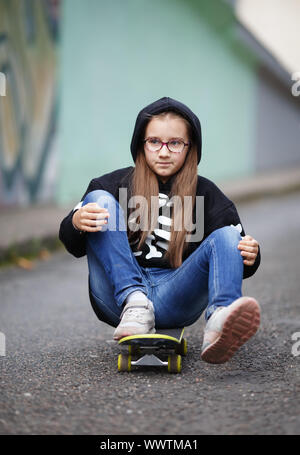 La ragazza corse su skateboard. Bambino in una felpa nera seduta sul suo skateboard. Colpo verticale. Messa a fuoco selettiva. Foto Stock