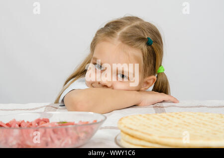 Bambina seduti ad un tavolo con la sua testa sulla sua mano con un sorriso e guardando il cibo di fronte a lei Foto Stock