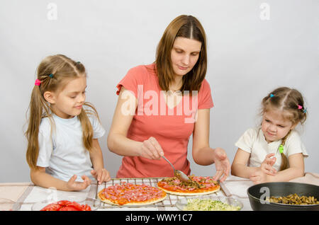 Due bambine seduto al tavolo della cucina e guardare come una madre preparare una pizza Foto Stock