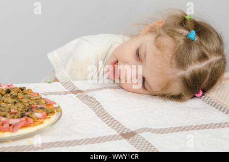 Bambina con linguetta sporgente appoggiata la testa sul tavolo e guarda la pizza Foto Stock