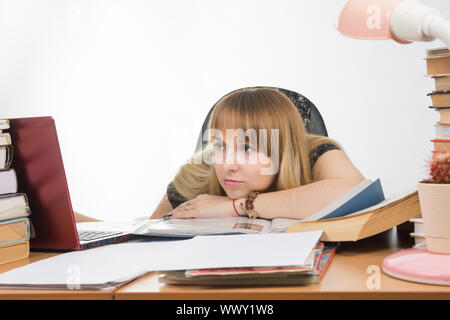 La ragazza alla reception stipate di libri e giornali purtroppo guarda il portatile con la sua testa in appoggio sulla sua mano sul tavolo Foto Stock