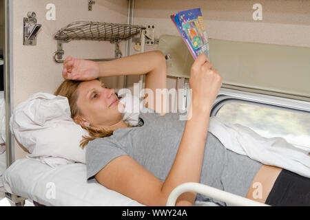 La ragazza sul treno stare sdraiato sulla schiena sul ripiano di sommità e di lato con un sorriso leggendo una rivista Foto Stock