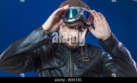 Pilota ingranaggio del 20s con occhiali da sole e vintage aviatore casco. Indossa giacca di pelle, barba e facce espressive Foto Stock