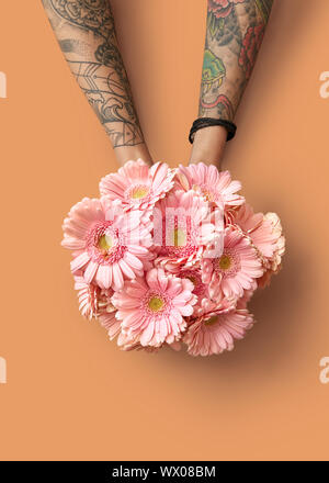 Le mani di una donna con un tatuaggio di tenere un bel bouquet con gerbere rosa su uno sfondo arancione Foto Stock