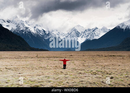 Uomo in cappotto rosso stava tenendo le braccia guardando delle montagne innevate, Parco Nazionale di Fiordland, UNESCO, South Island, in Nuova Zelanda, Pacific Foto Stock