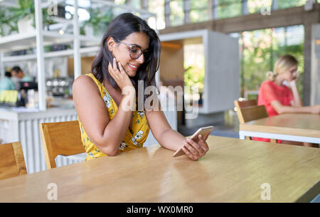 Candide lifestyle ritratto di sorridente millenaria ispanica designer di moda di scattare una foto sul cellulare in una moderna e luminosa cafe con piante Foto Stock